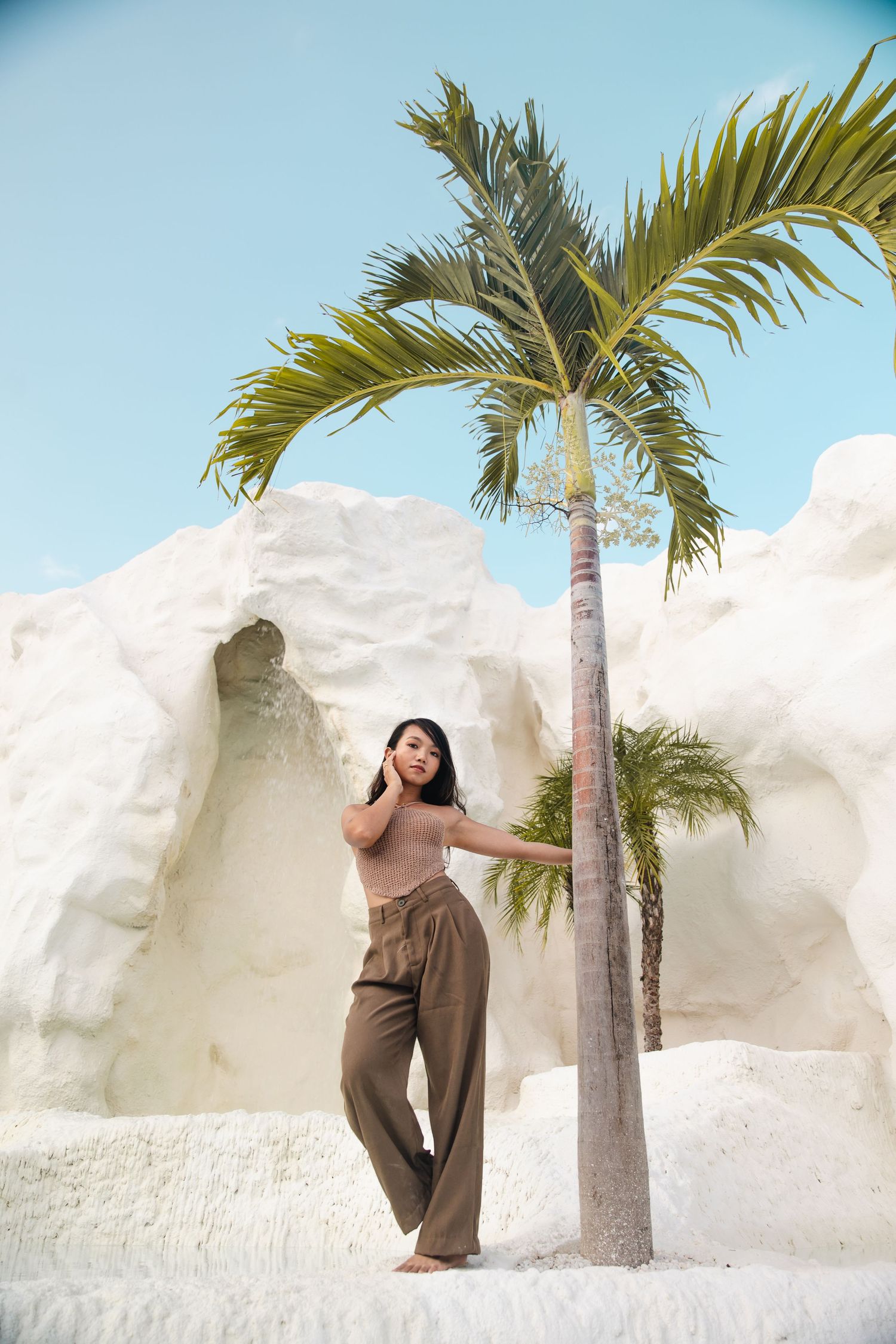 A woman in casual attire poses against white walls beside a tall palm tree on a sunny day.