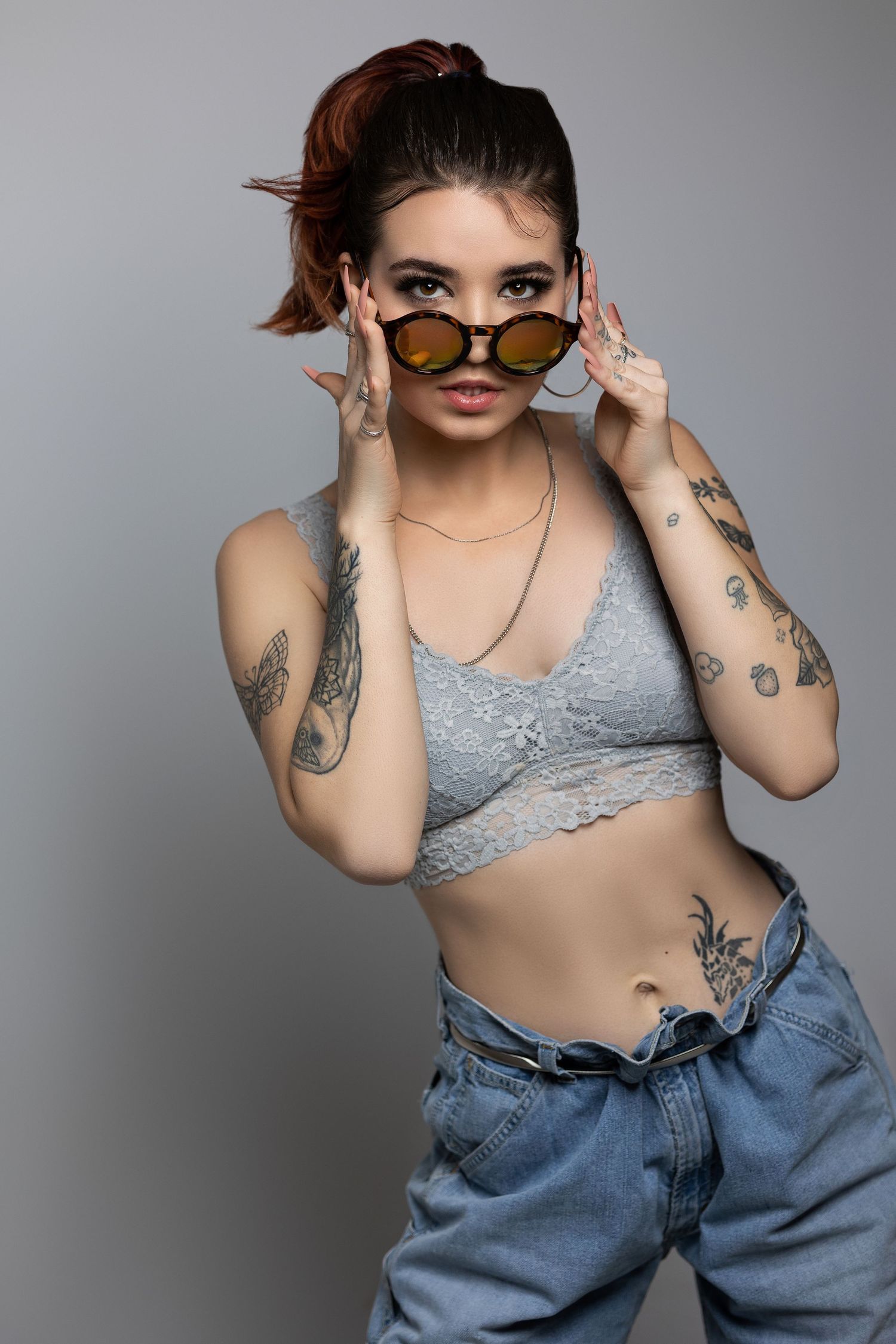 Alternative style portrait with sunglasses, tattoos, and crop top against grey background.