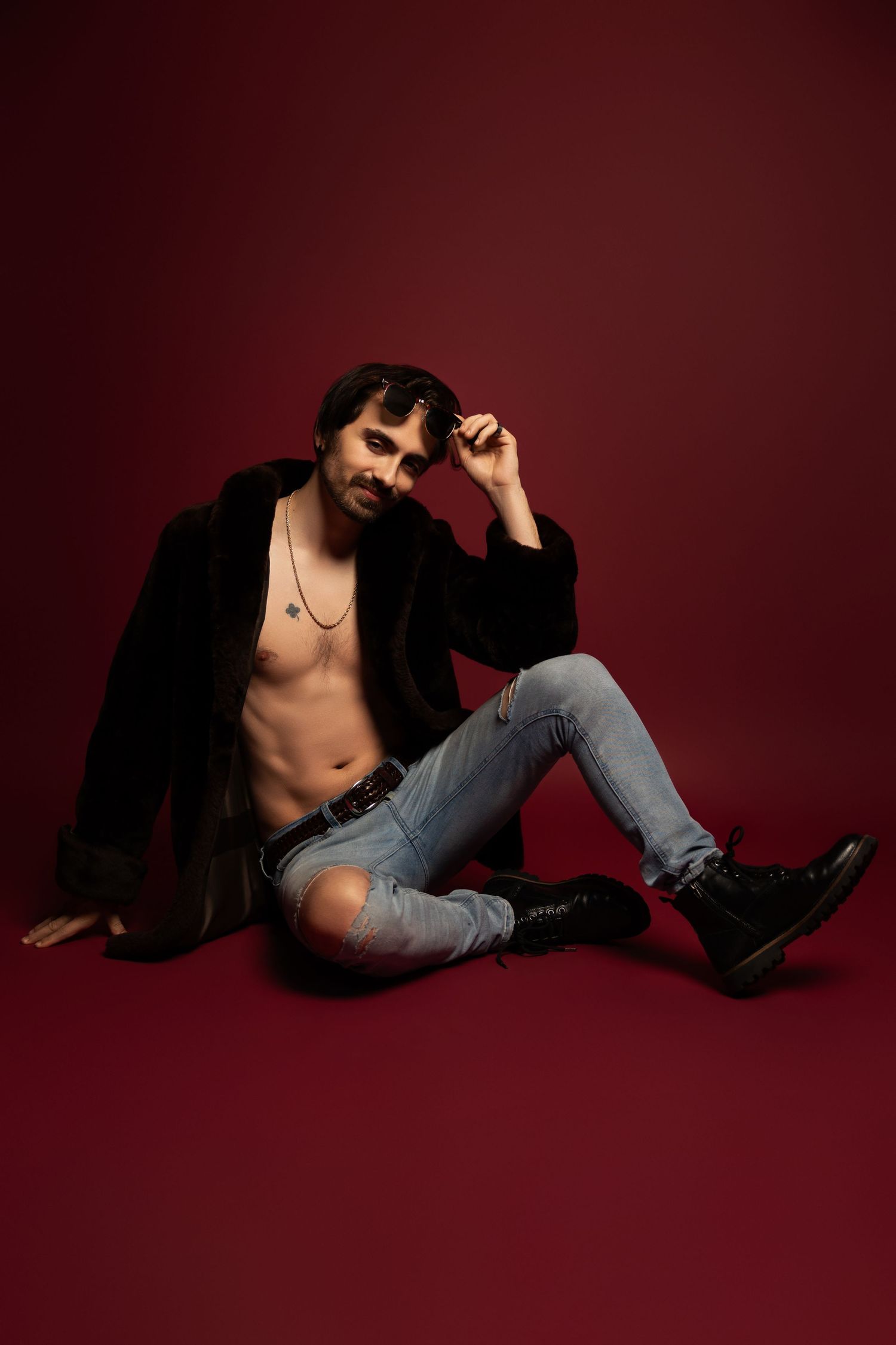 Fashion model in ripped jeans and black jacket poses against dark red background in artistic portrait session.