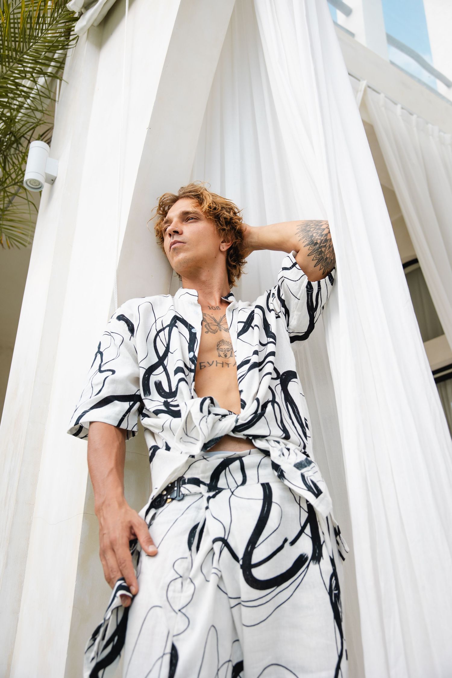 Figure in black and white abstract patterned outfit poses against white columns in tropical setting.