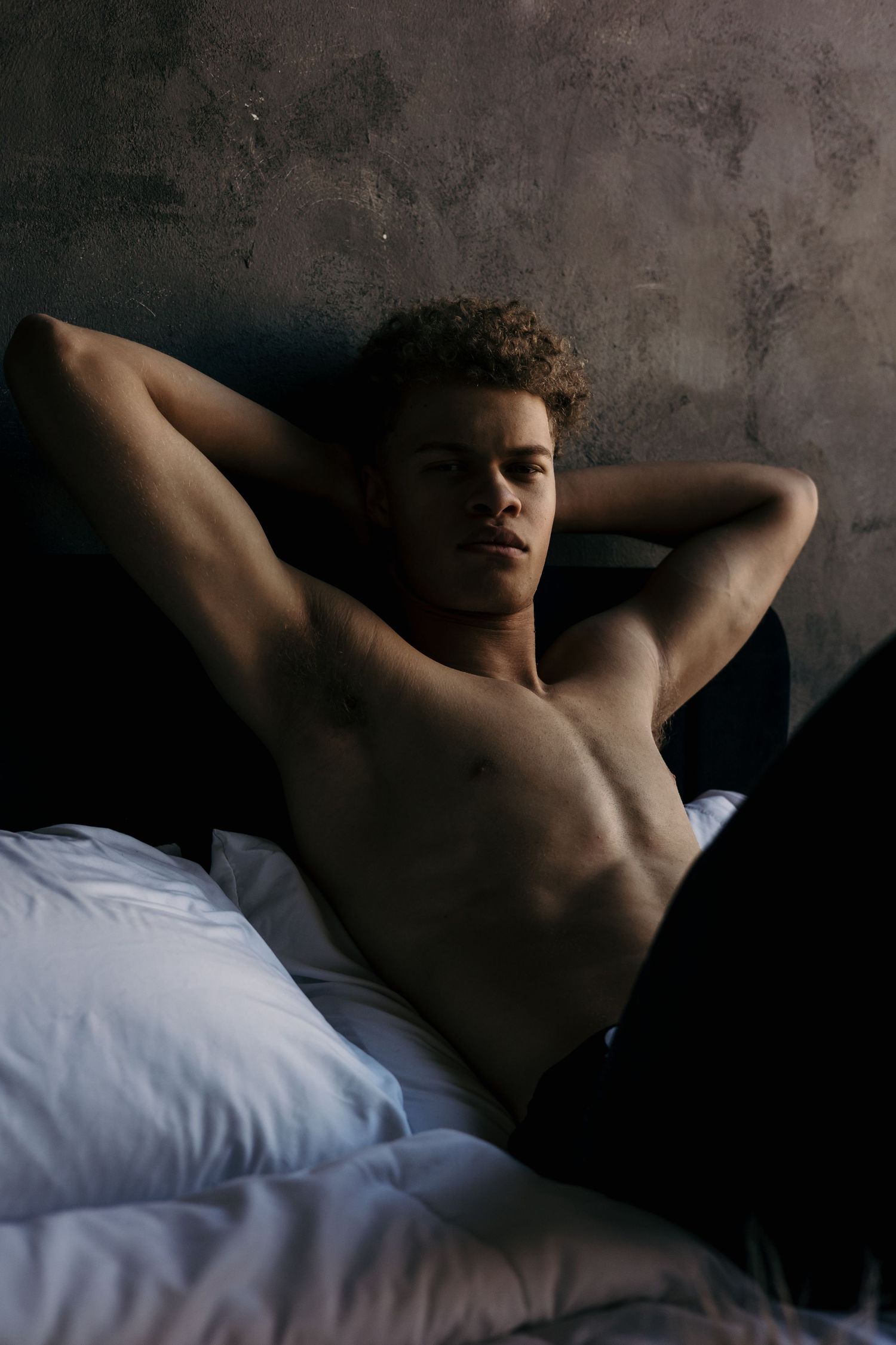 Person reclining on bed in moody lighting against textured wall.