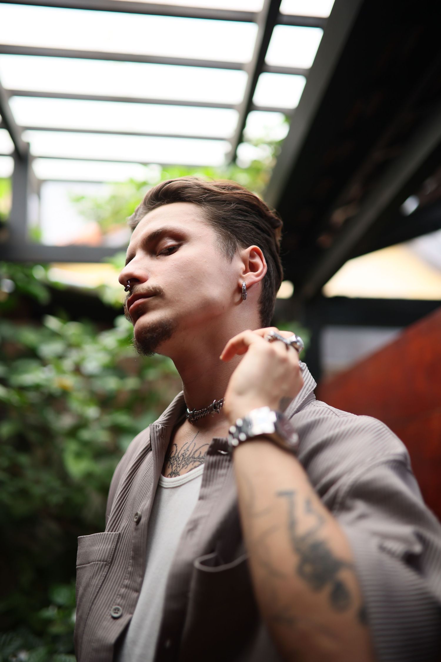 Stylish person in grey button-up shirt with tattoos and jewelry posing thoughtfully in outdoor covered space.