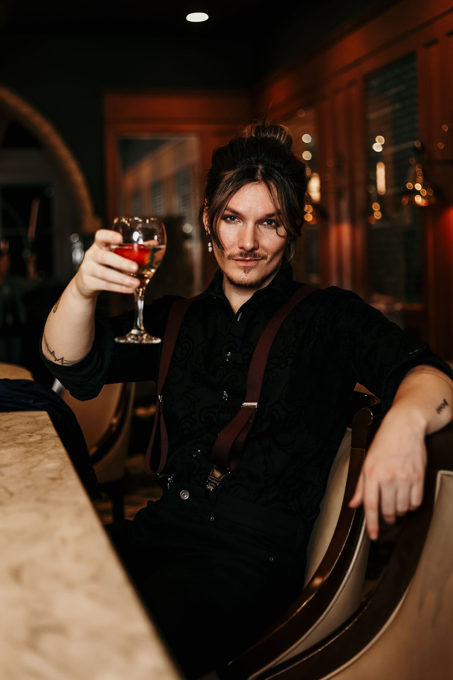 Person in black attire with suspenders toasting with wine glass in dimly lit bar setting.