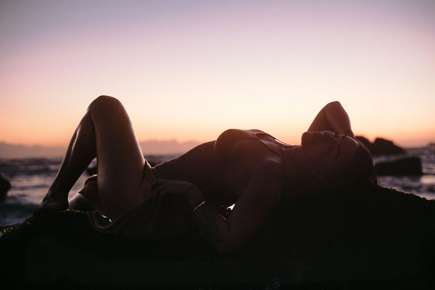 Silhouetted figure resting on rocks at sunset by the ocean shore.