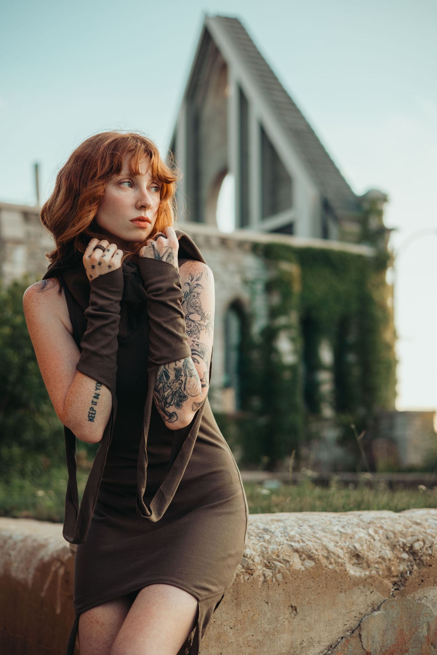 Brown dress and arm warmers with tattoos against architectural backdrop in moody portrait photography.