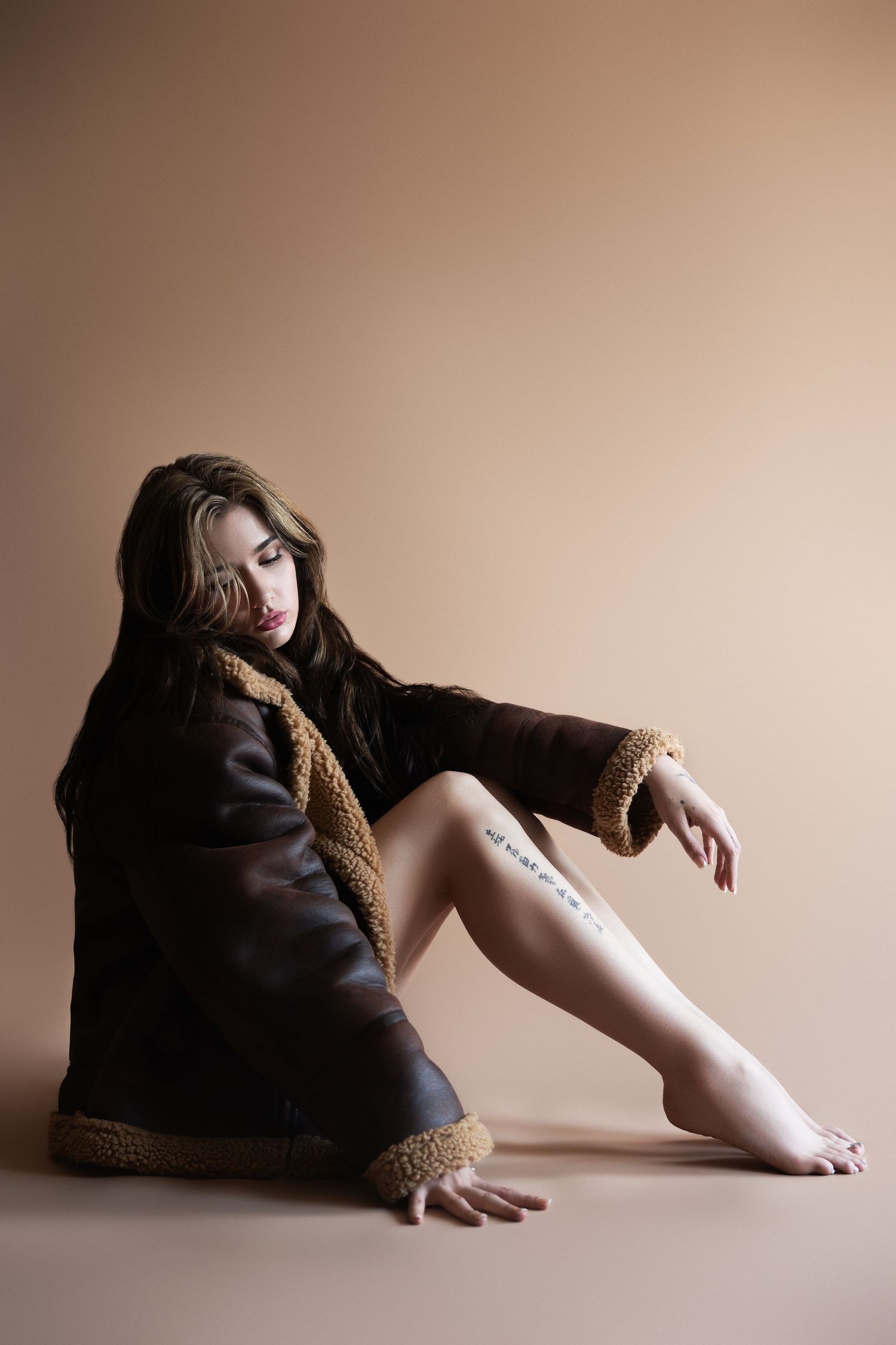 Artistic portrait of someone wearing a brown shearling coat sitting against beige background with leg tattoo visible.