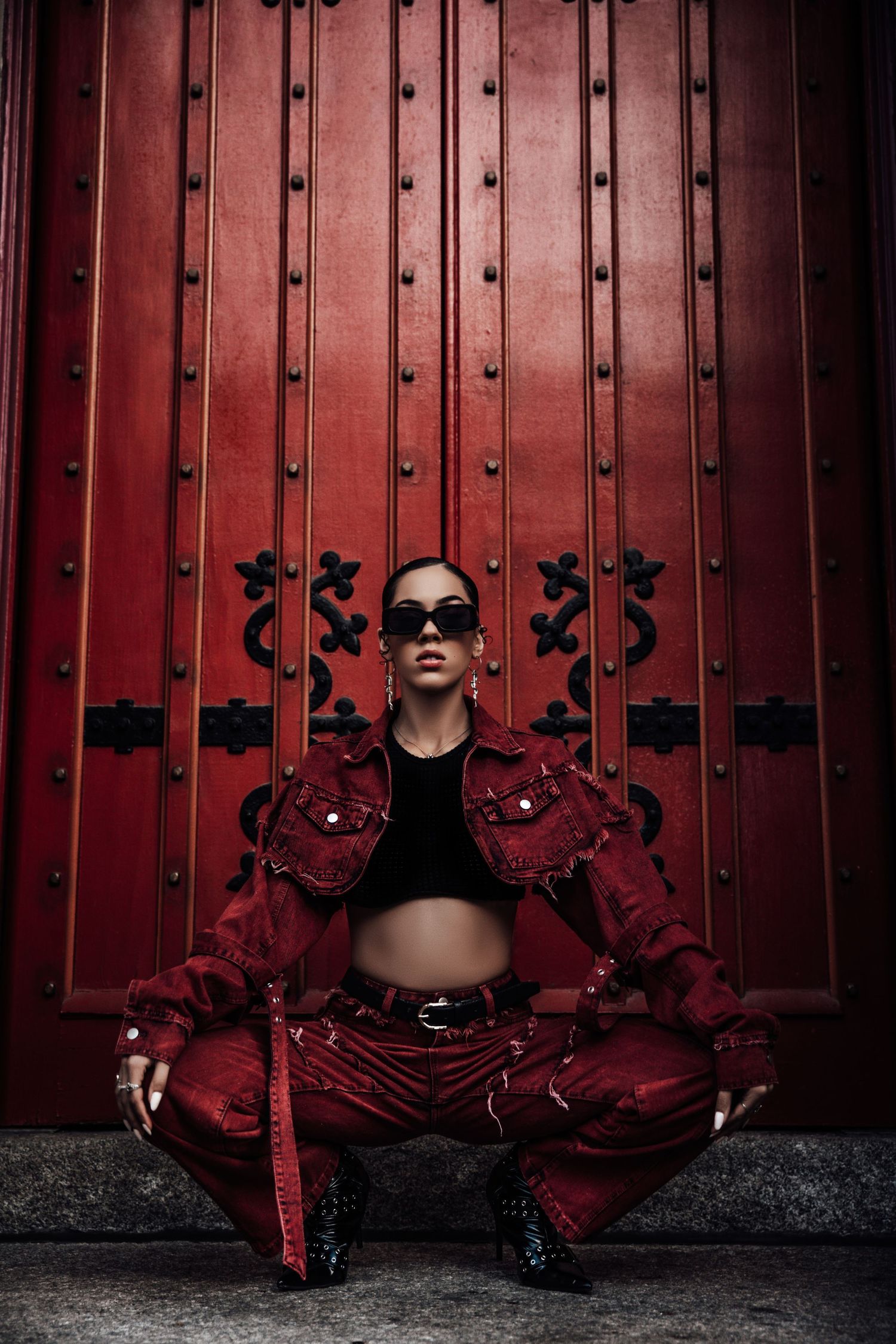 Stylish figure in red outfit and sunglasses poses in front of ornate burgundy doors with decorative metalwork.