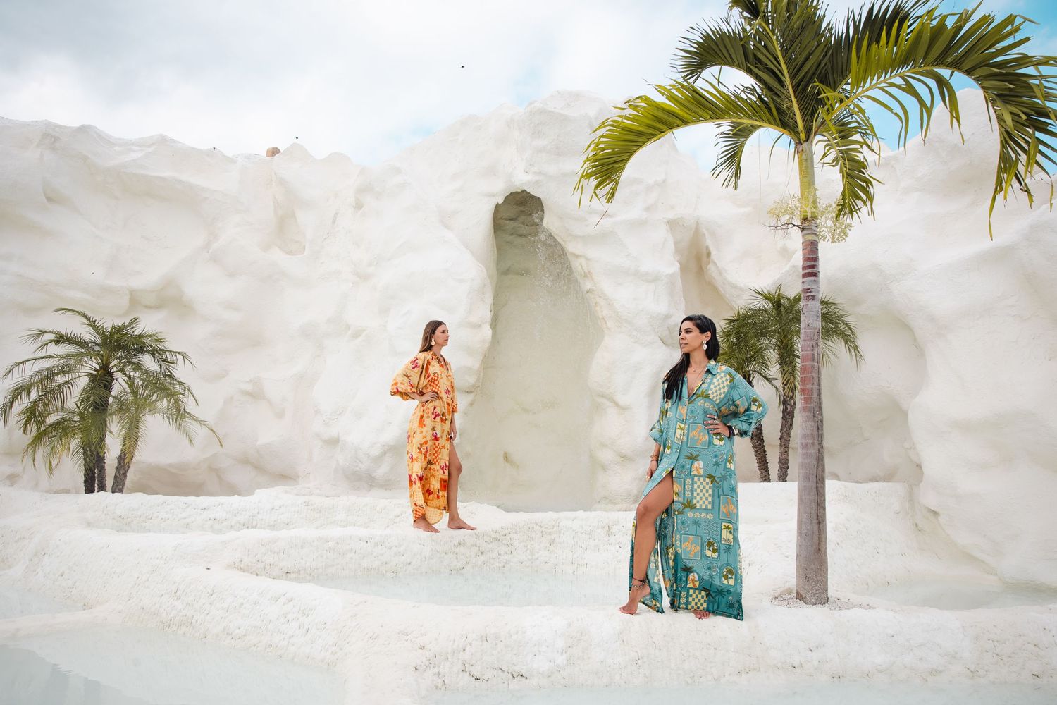 Two people in colorful maxi dresses posing against white walls with tropical palm trees.