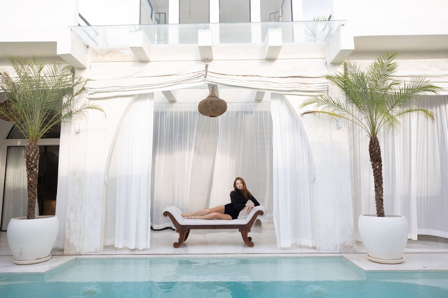 Elegant poolside cabana with white curtains and palm trees features a comfortable lounge chair by a tranquil swimming pool.
