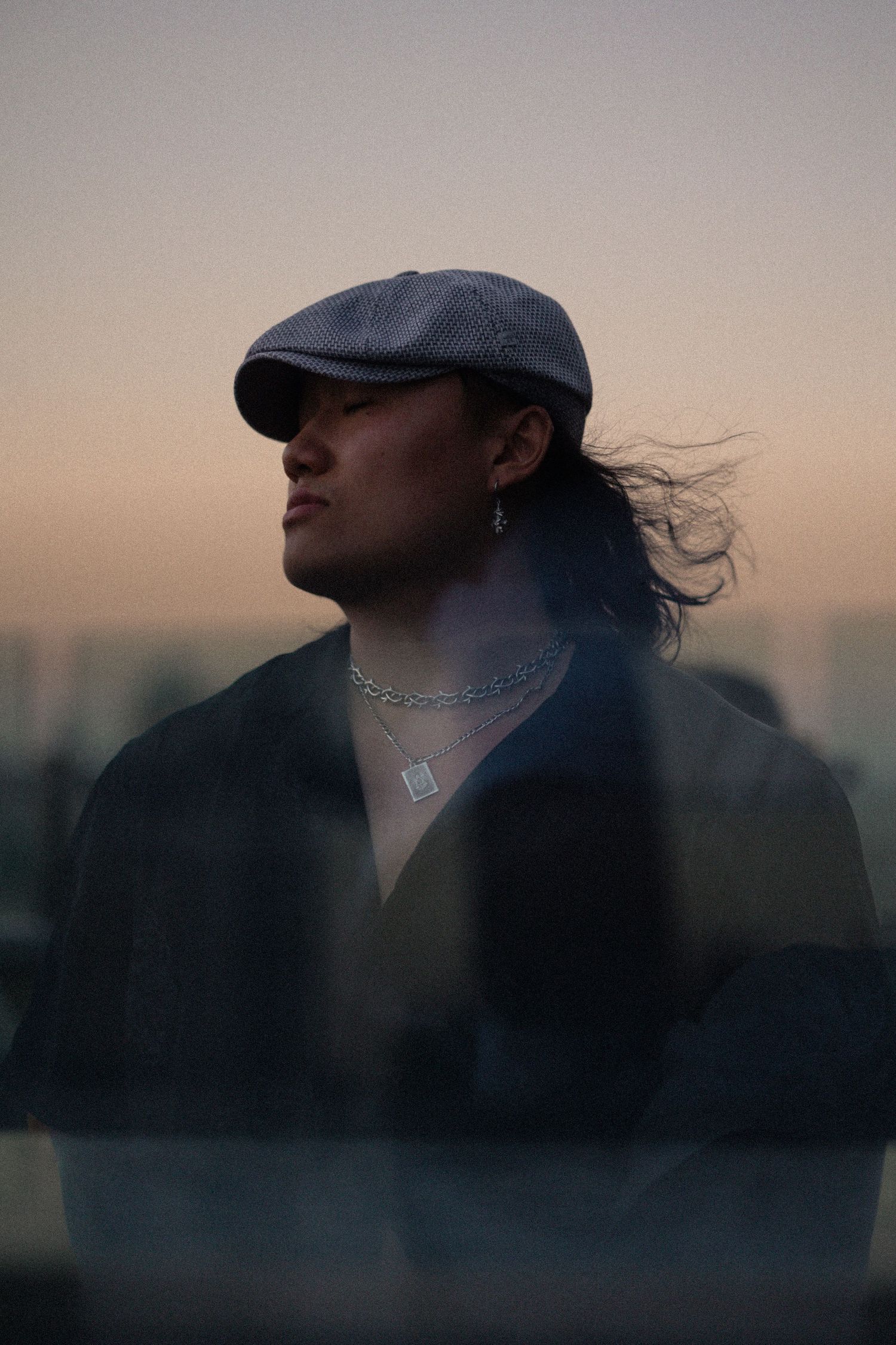 Silhouette wearing flat cap and jewelry against sunset sky with moody lighting.