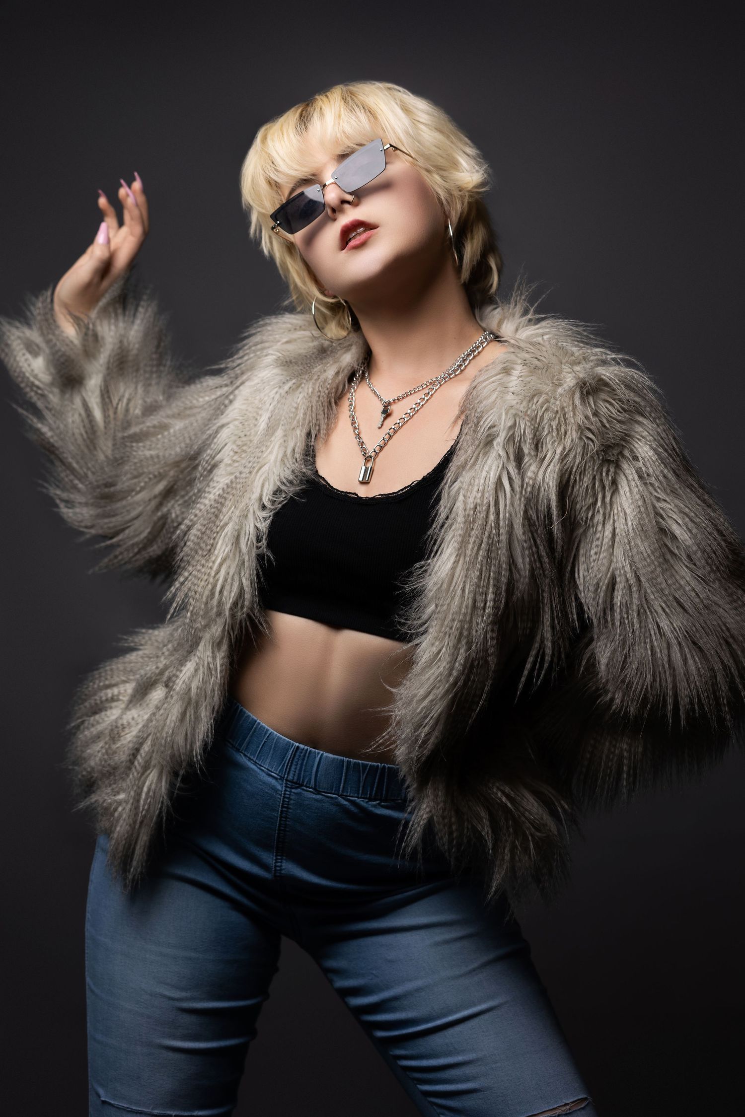 Fashion model in fur coat, crop top and jeans striking dramatic pose against dark background with sunglasses.