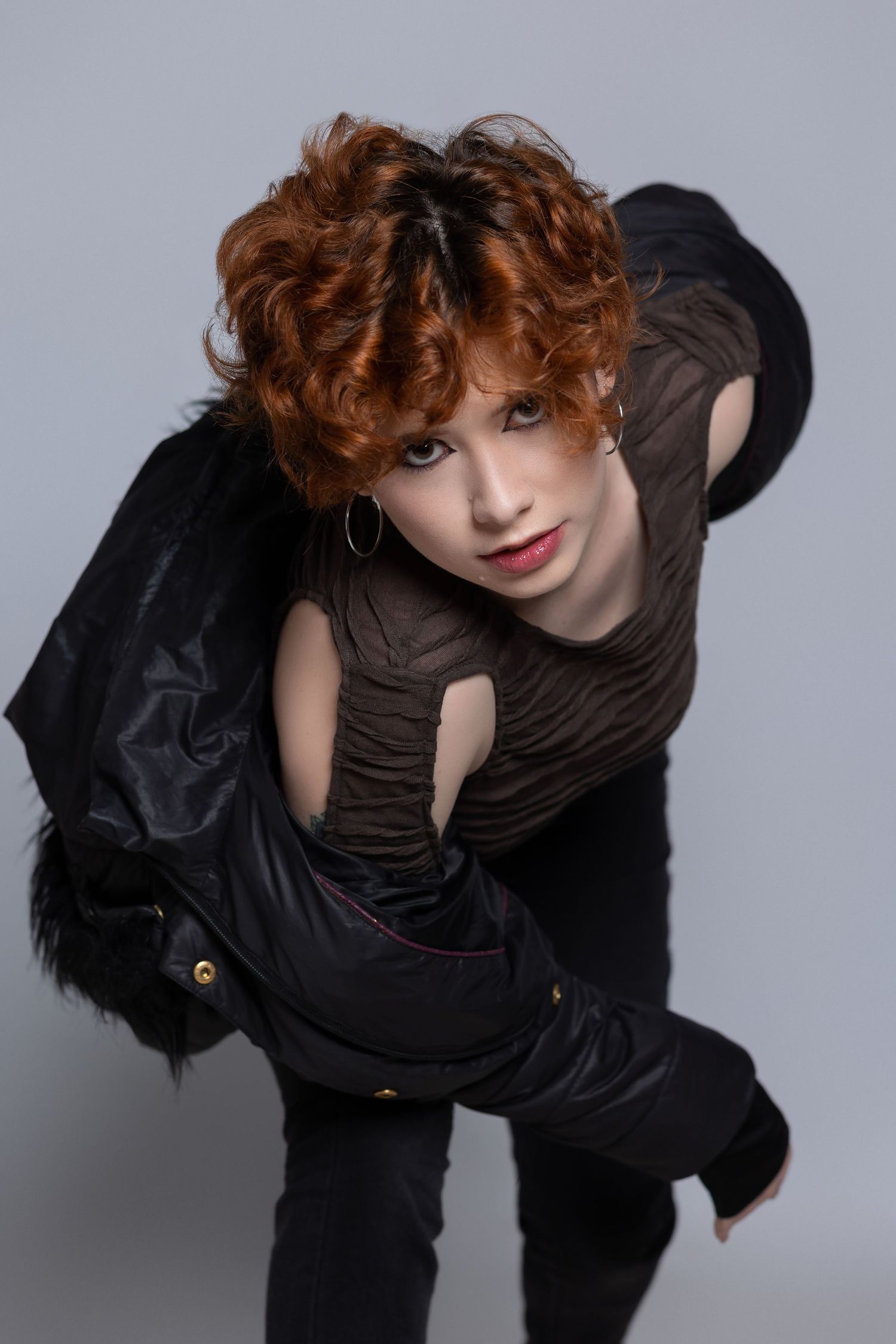 High-fashion aesthetic studio portrait with dramatic pose wearing black clothing and vibrant red curly hair against gray backdrop.