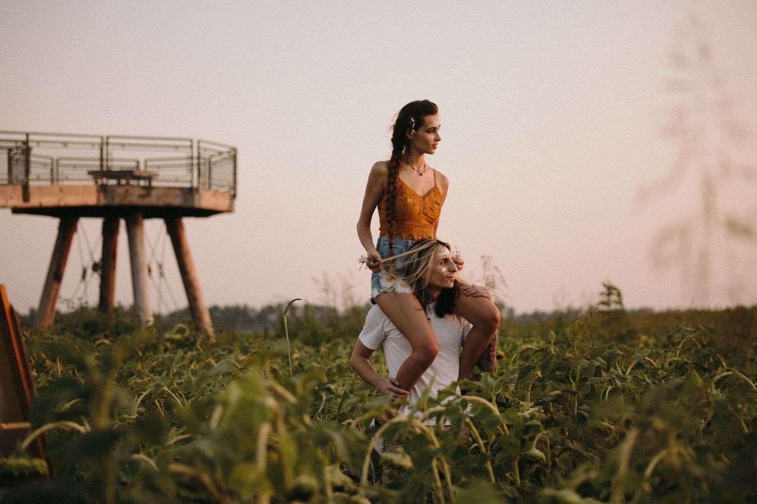 Person in orange tank top and denim shorts posing near industrial structure in field at dusk.