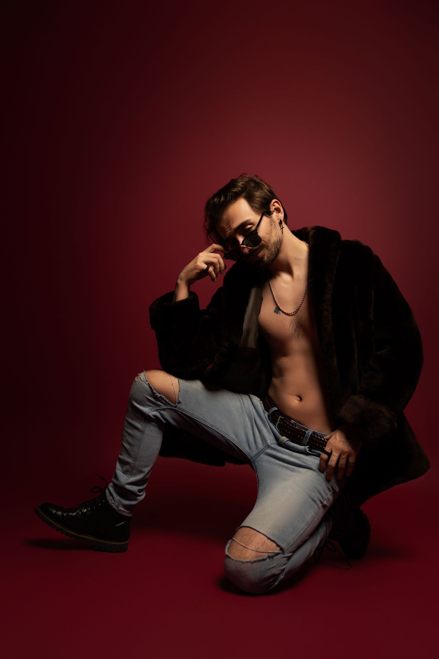 A model poses in a black fur coat and ripped jeans against a dark red backdrop in dramatic studio lighting.