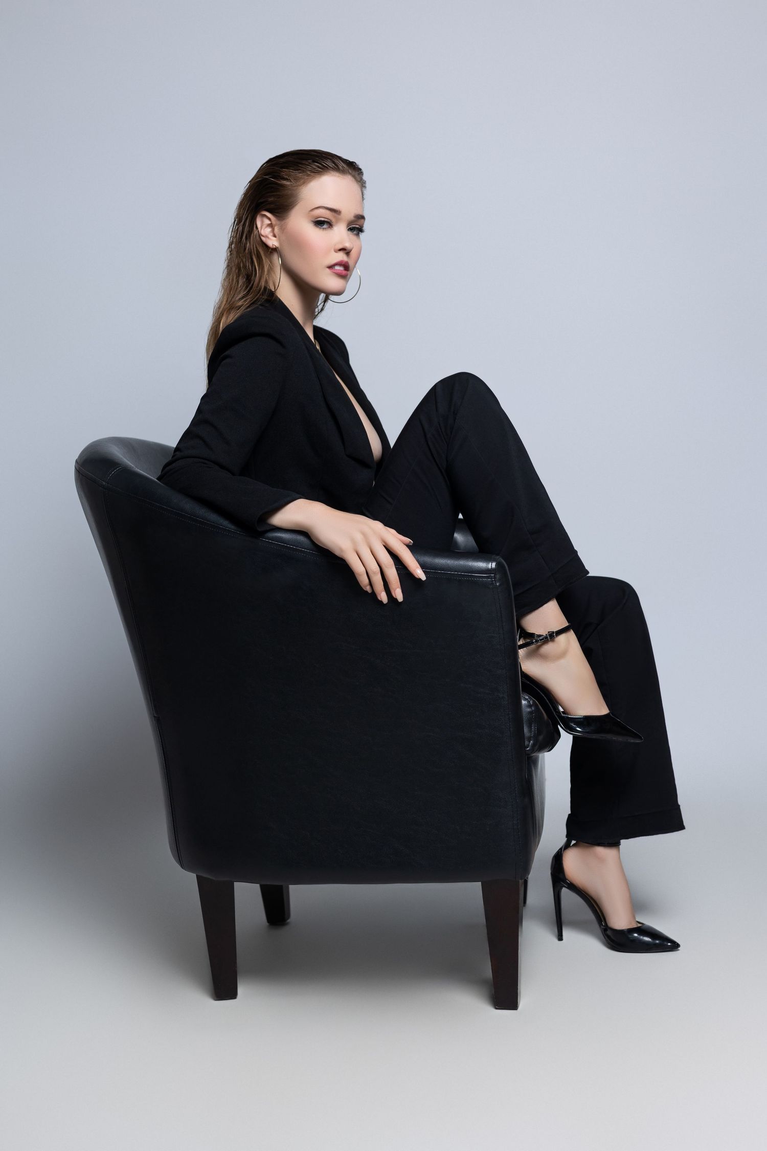 Fashion model in black pantsuit and heels poses elegantly in dark armchair against gray studio background.