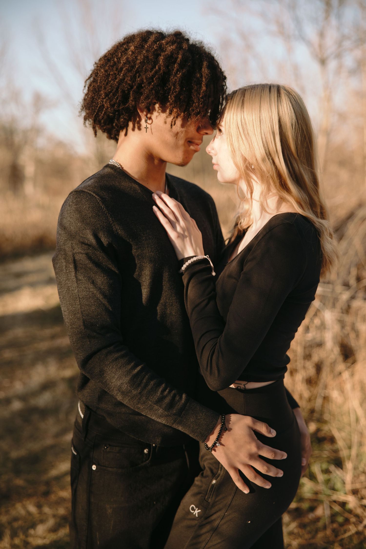 Intimate couple in dark clothing embracing against natural winter background.