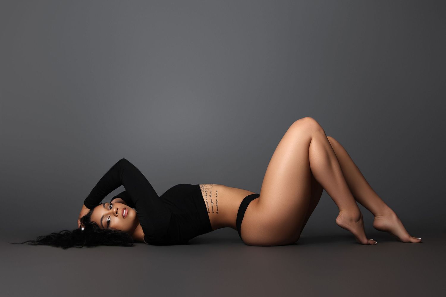 Model in black outfit lying on dark floor during artistic photoshoot.