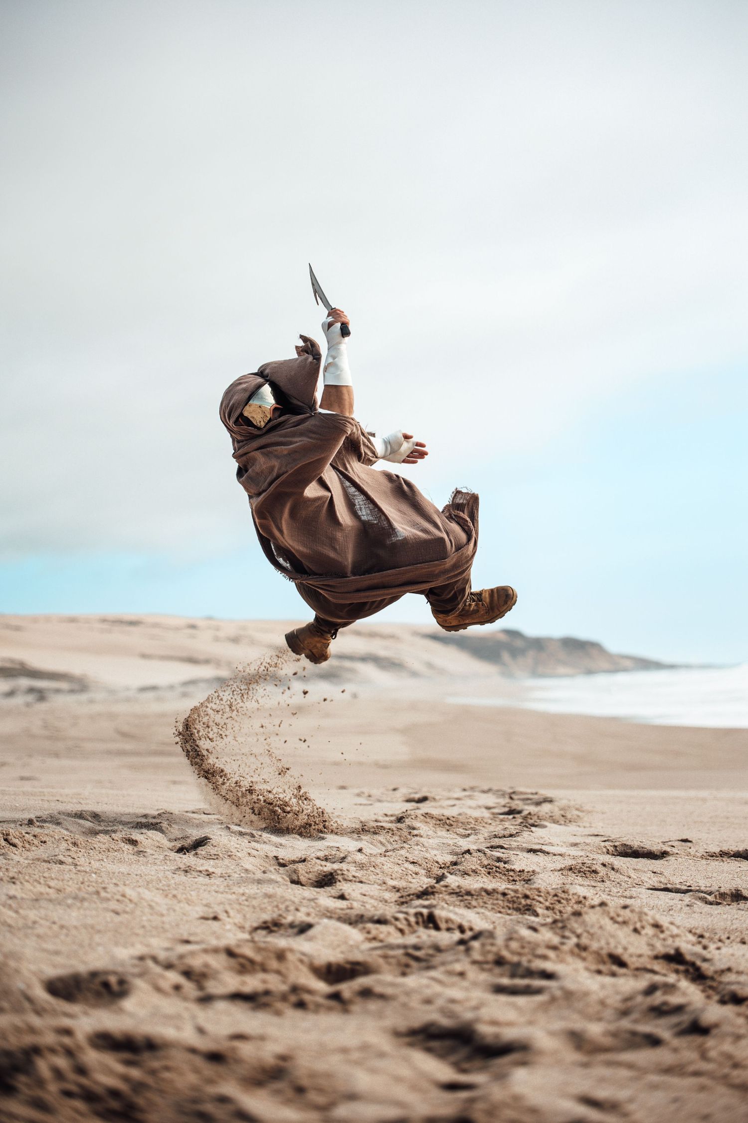 Figure in brown flowing robes leaps dramatically over desert sand, kicking up particles against a bright sky.