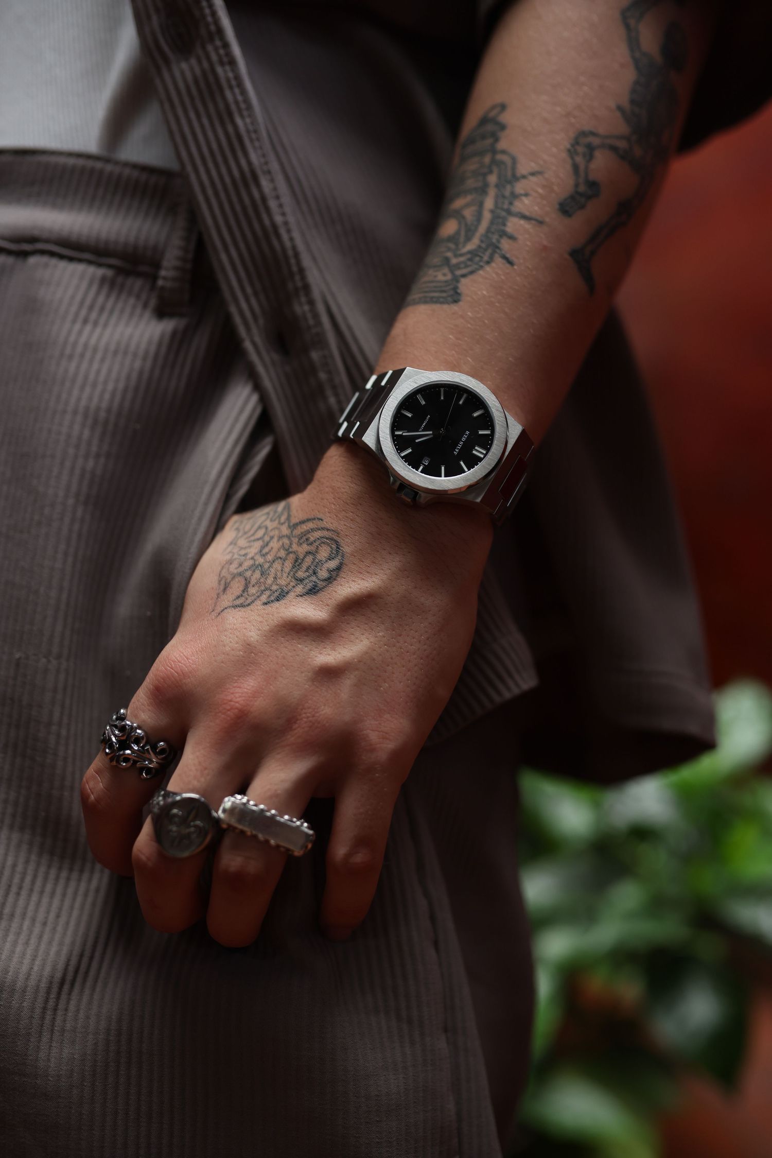 Close-up of tattooed arm wearing silver watch and multiple rings against brown clothing background.