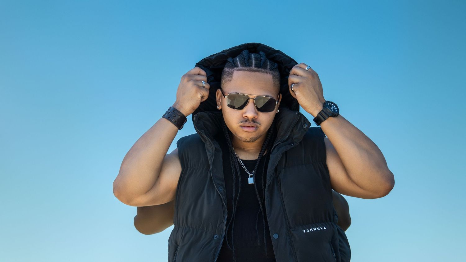 A person in sunglasses and a black hooded vest adjusts their hood against a bright blue sky background.