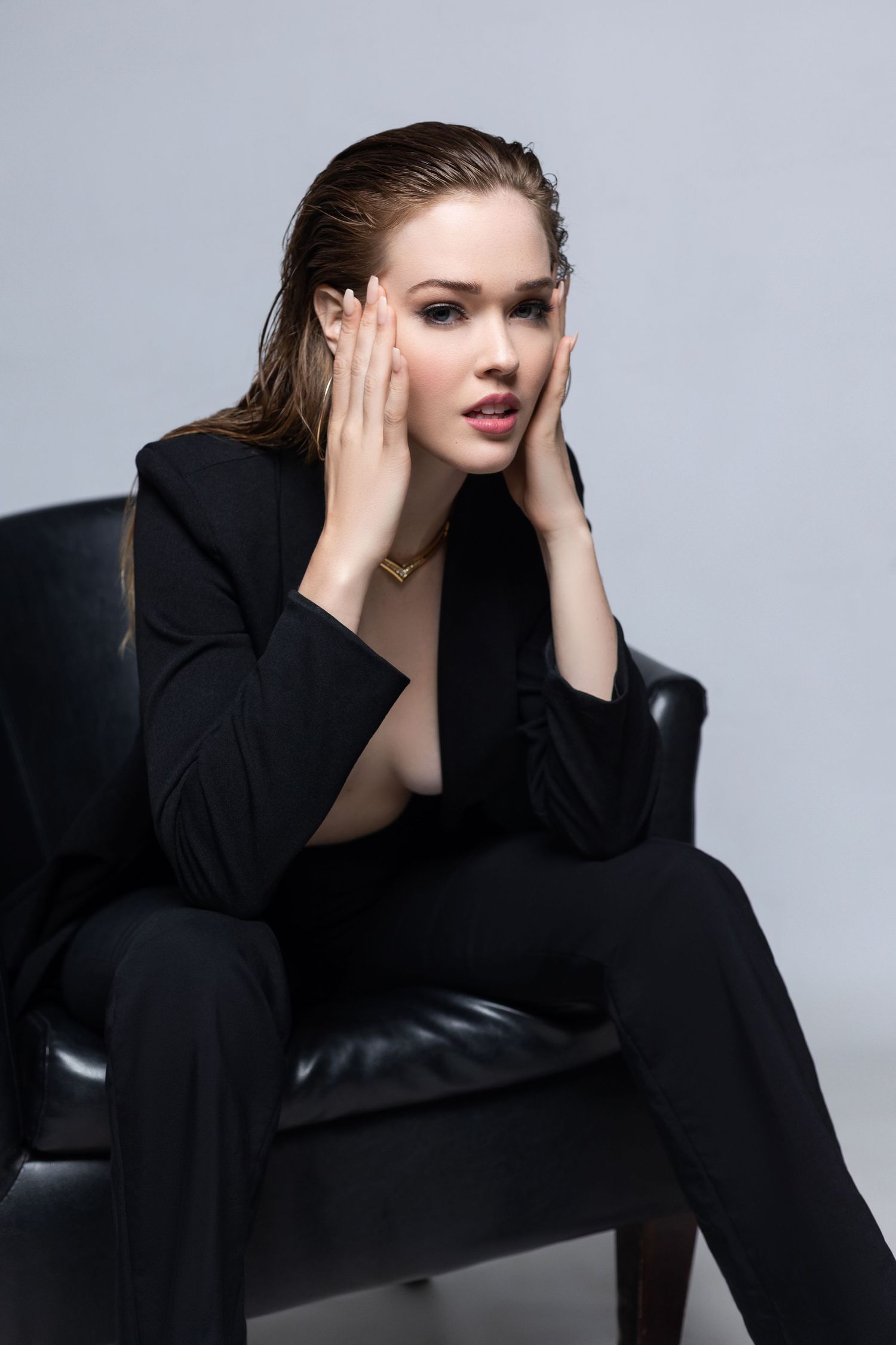 Fashion portrait of model in black suit jacket sitting on leather armchair against gray backdrop.