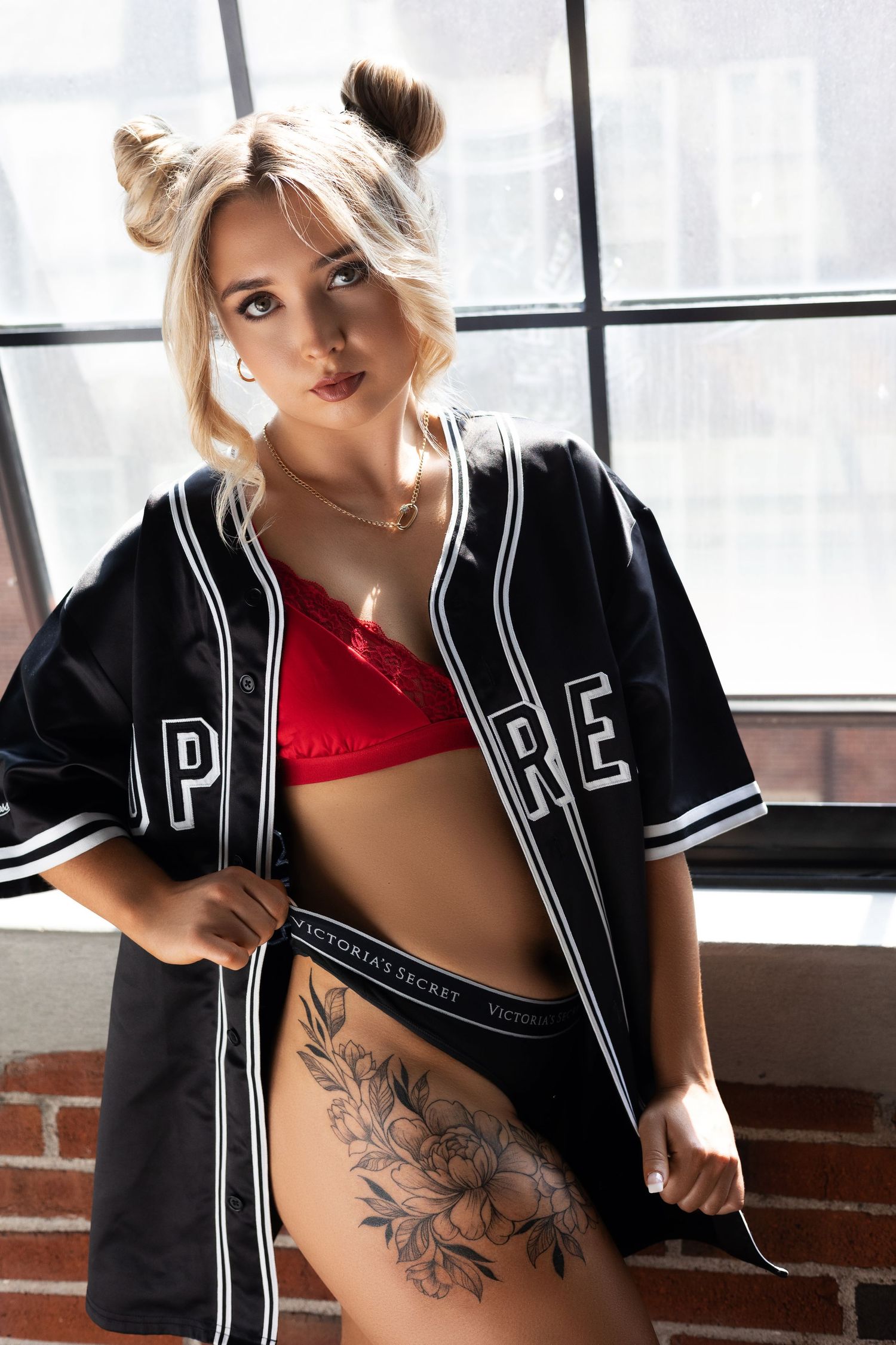 Athletic fashion look featuring black varsity jacket, red bralette and floral hip tattoo near window.