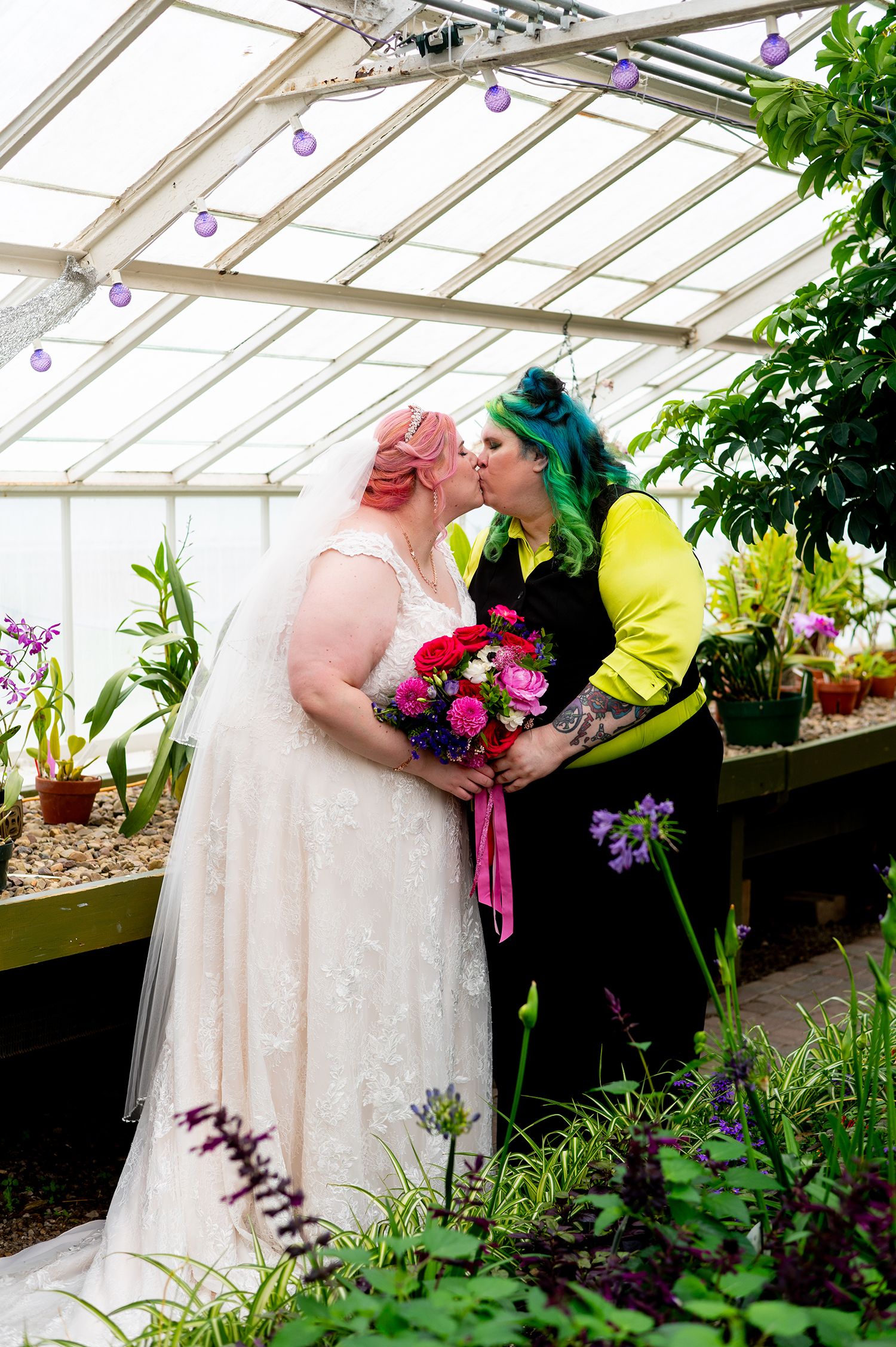 Two brides sharing a kiss in a greenhouse, one in a white lace gown and veil, the other in a black and yellow outfit.