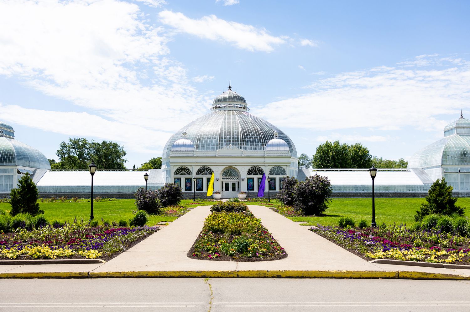 Grand glass dome greenhouse botanical garden with manicured flower gardens and tree-lined pathway on a sunny day.