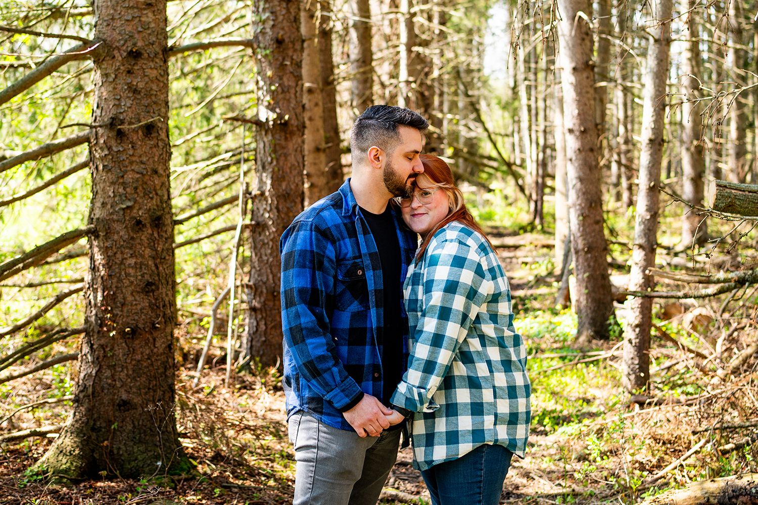 A couple in matching plaid flannel shirts embrace tenderly in a sunlit forest setting.