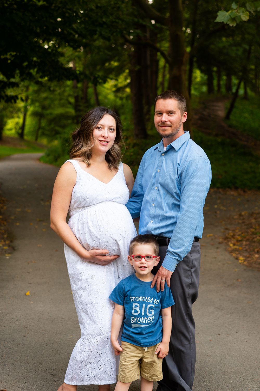 A family poses together on a wooded path, featuring a pregnant mother in white and a child in blue.