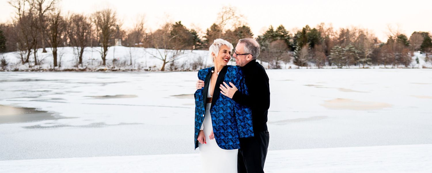 A loving couple embrace while standing in a snowy winter landscape at sunset with trees in the background.