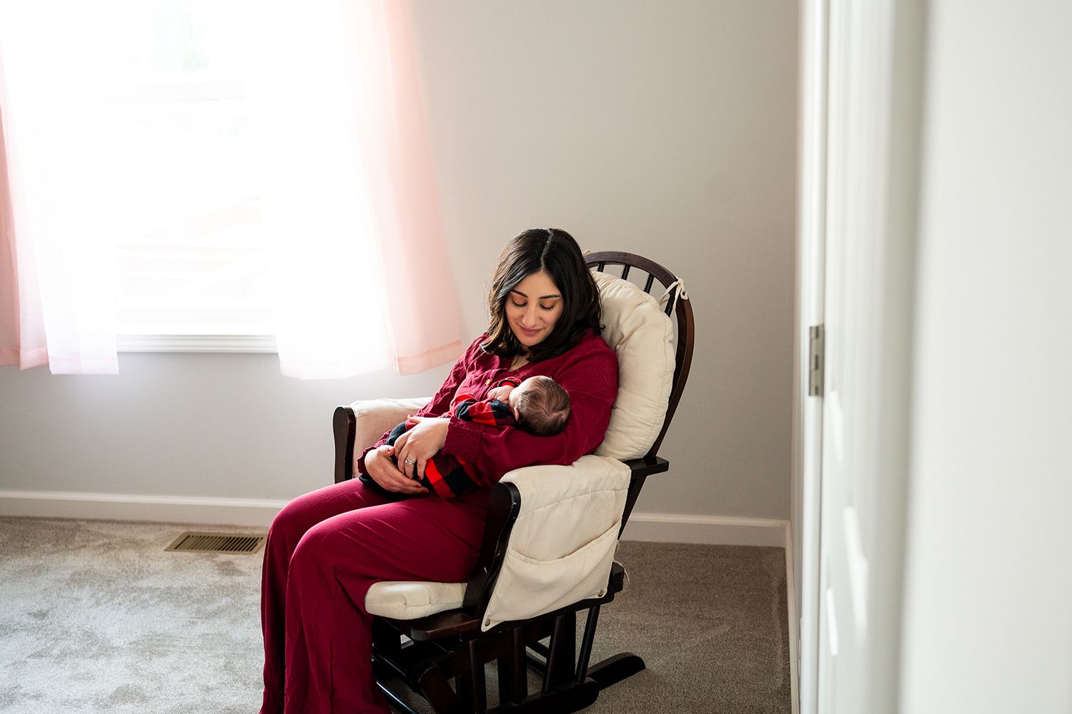 A person in red pajamas sits comfortably and relaxes in a nursery rocking chair next to a bright window.