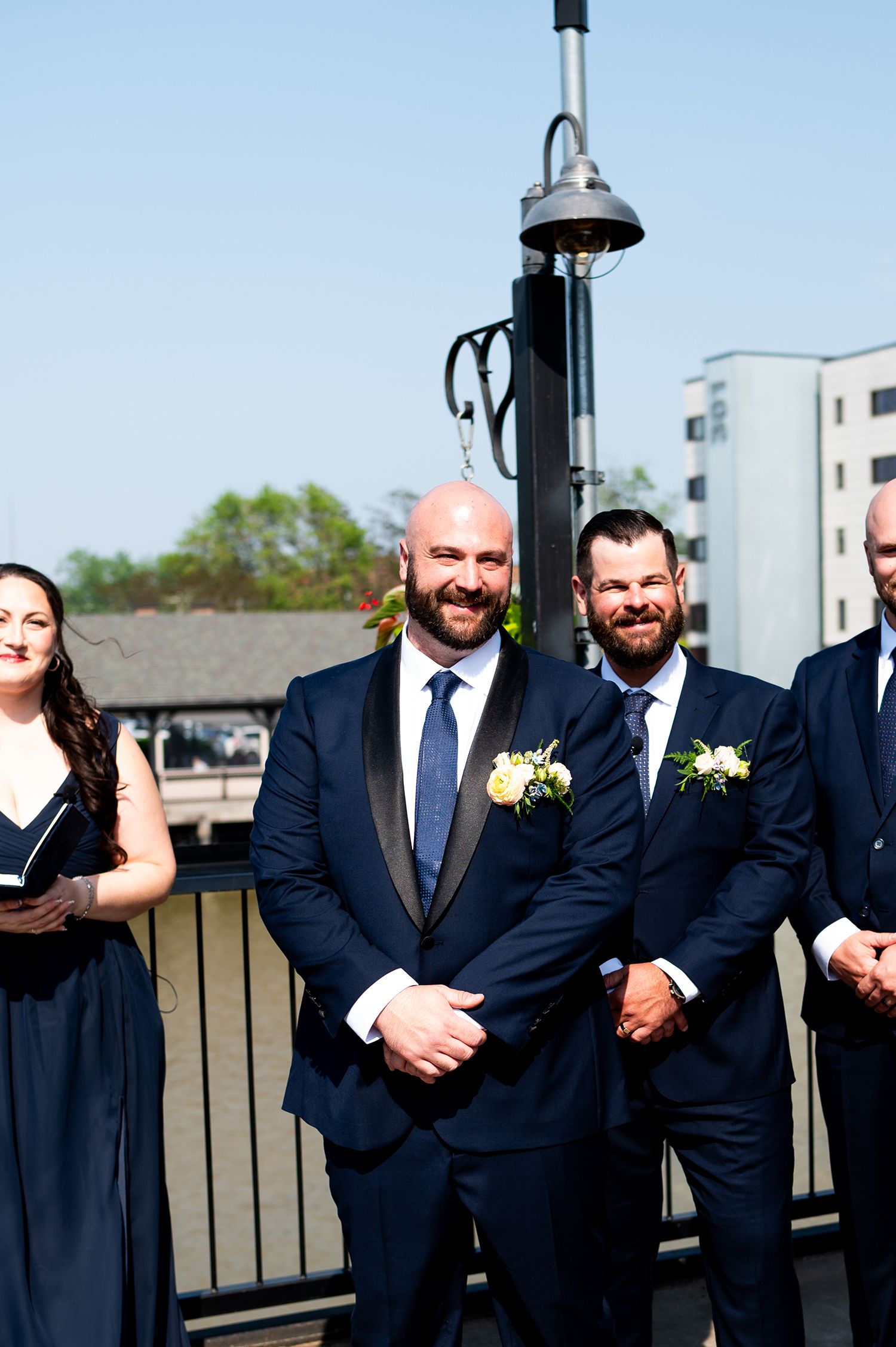 A group wearing navy suits and boutonnieres poses together outdoors on a rooftop terrace.