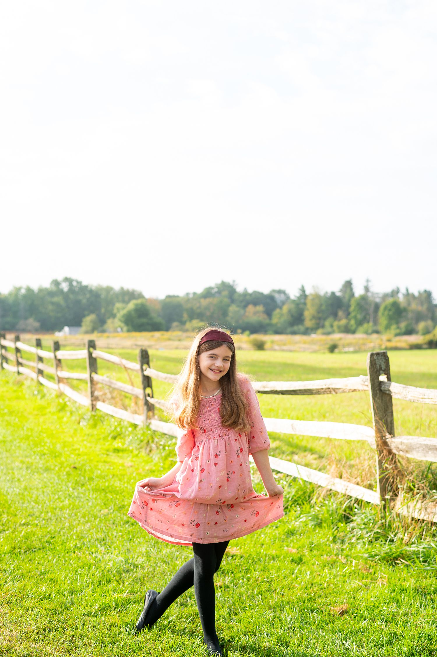 Young girl in pink dress and black tights twirling outdoors near a wooden fence on a sunny day.