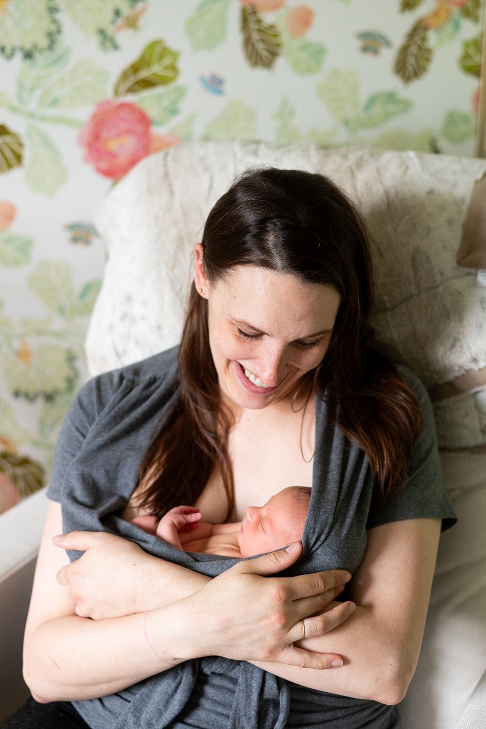 Woman breastfeeding newborn baby while sitting on chair with floral wallpaper background.