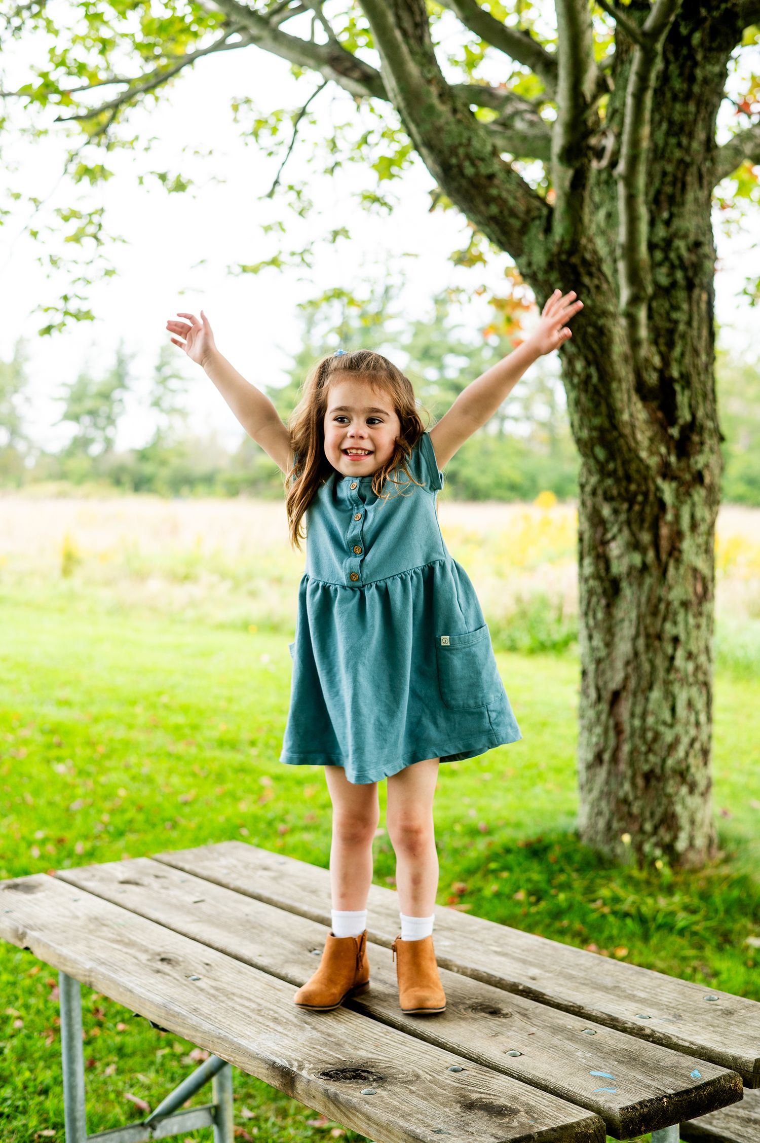 A joyful young girl in a teal dress stands on a picnic table with arms raised outdoors near a large tree.