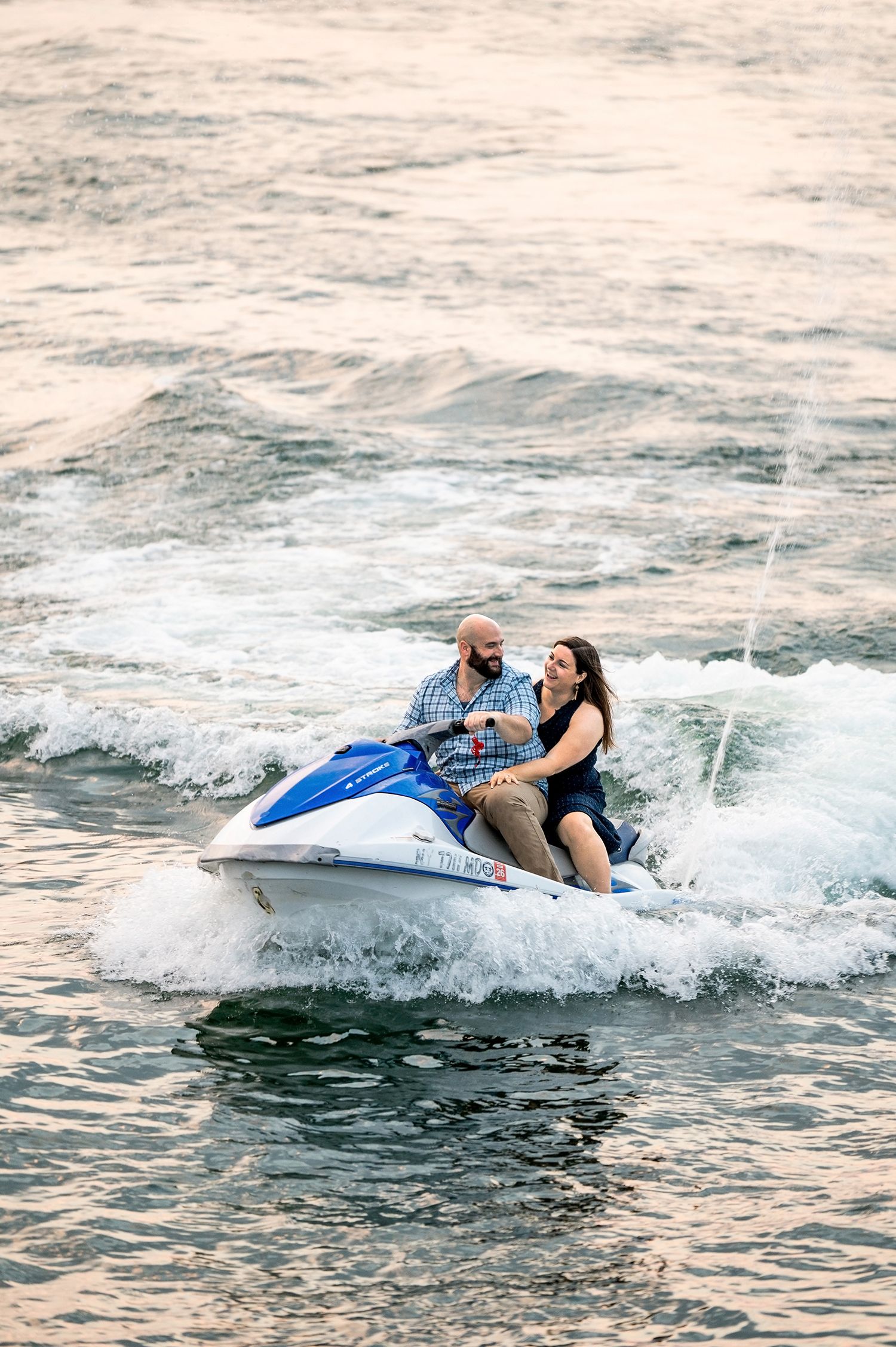 Couple smiling on a blue jet ski creating white water spray during a romantic sunset ride on the lake.