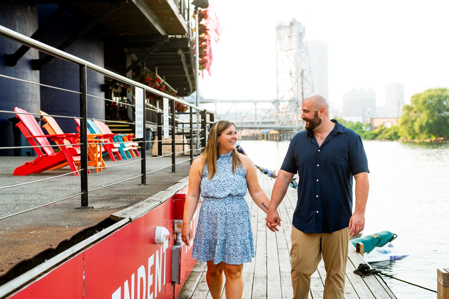 Couple holding hands and smiling on a waterfront dock with colorful chairs and city skyline in background.