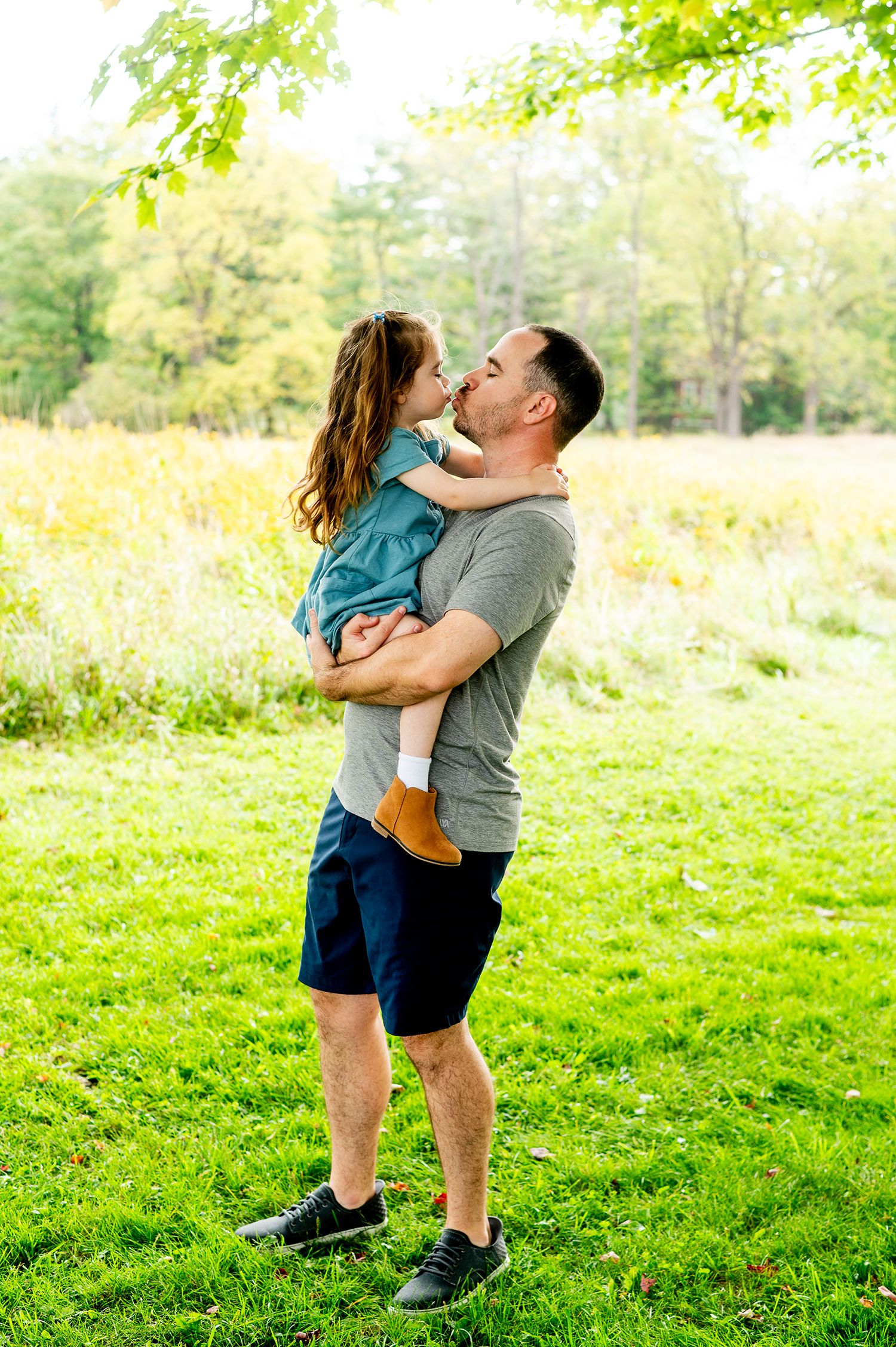 A father kisses his young daughter while holding her up in a sunny green park setting.