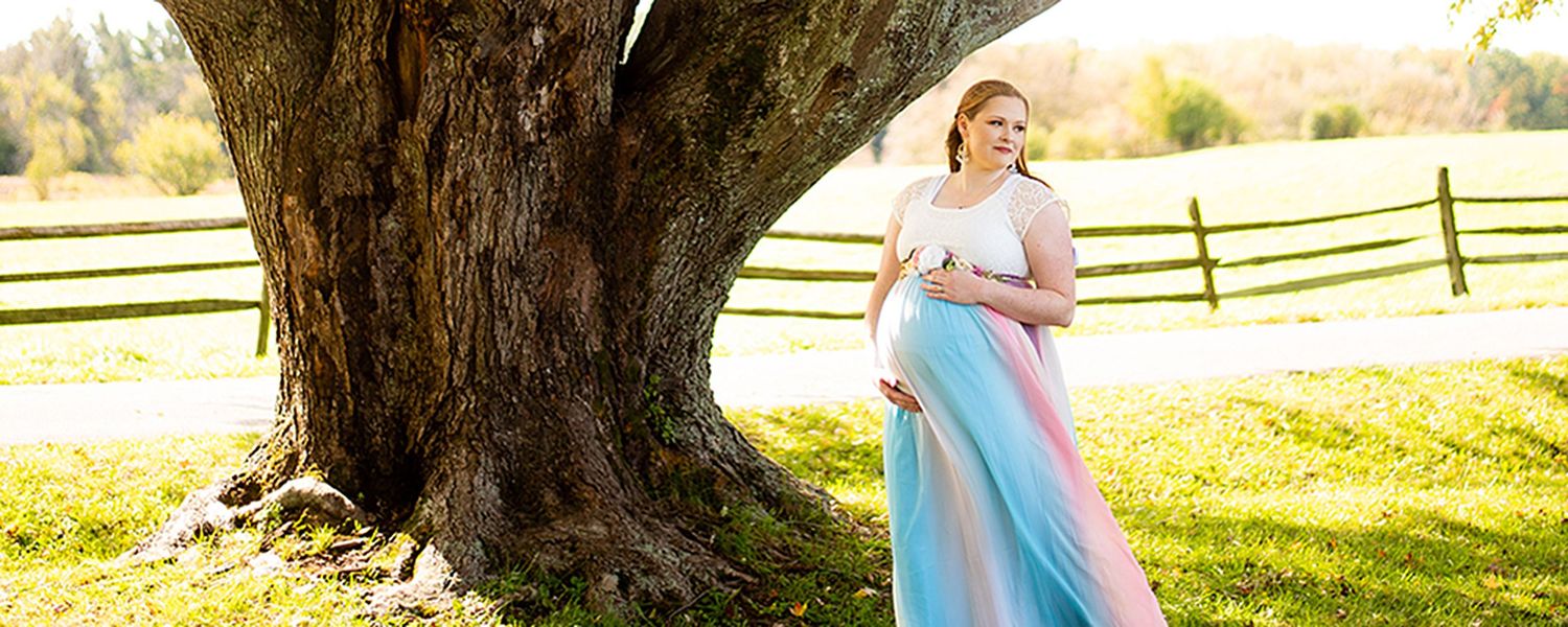 A maternity photo showing a woman in a flowing blue dress standing by a large tree trunk in a sunlit field.