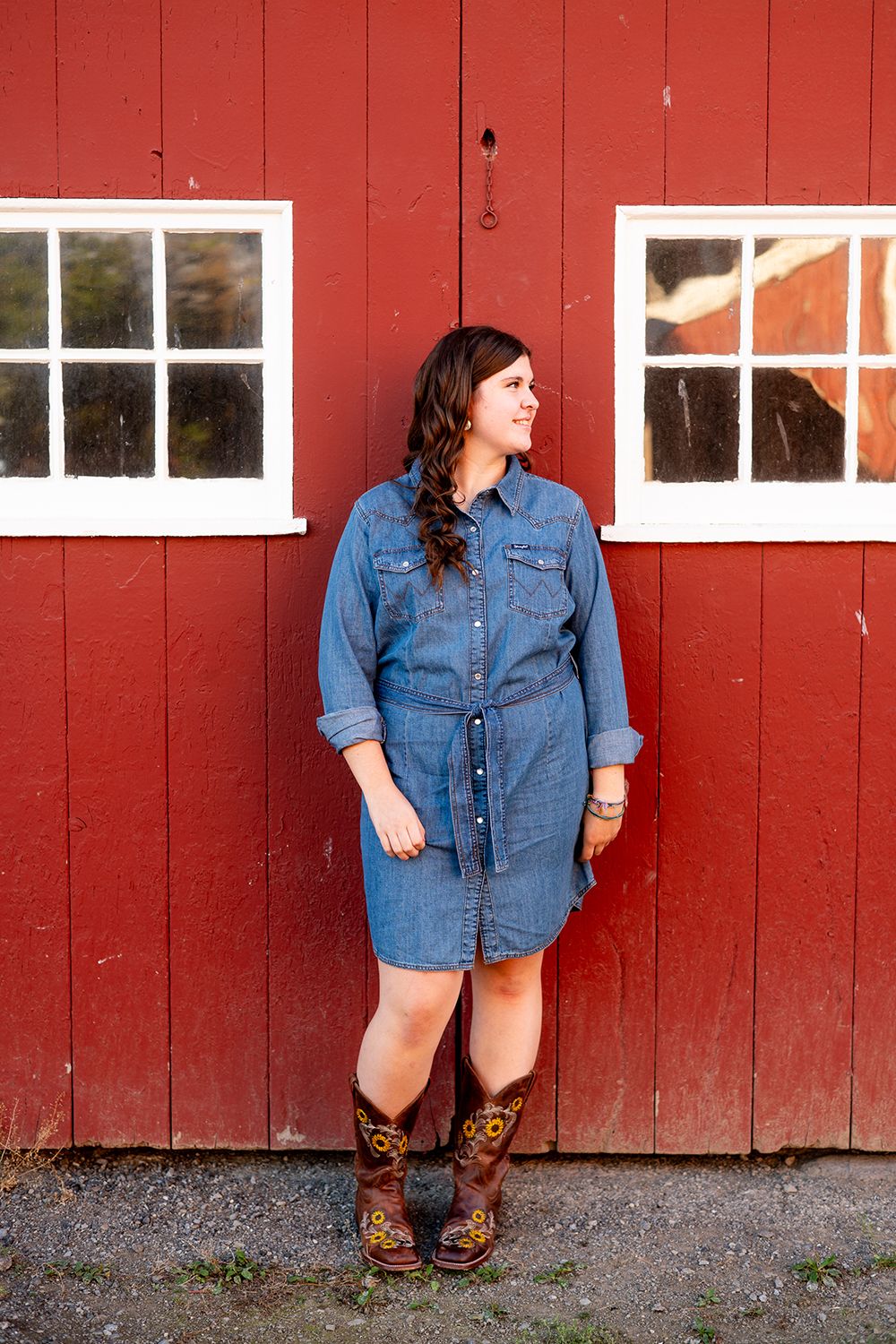 A person in a denim shirt dress and brown cowboy boots poses against a bright red barn wall.