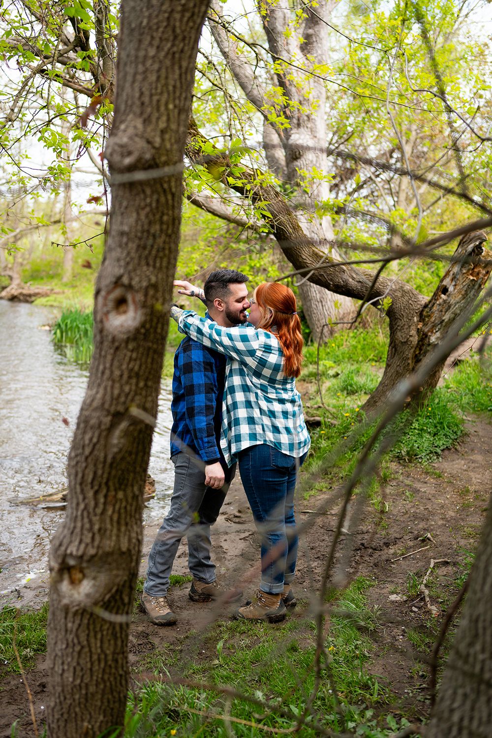A couple enjoys a quiet moment together on a trail by a stream, both wearing plaid shirts on a spring day.