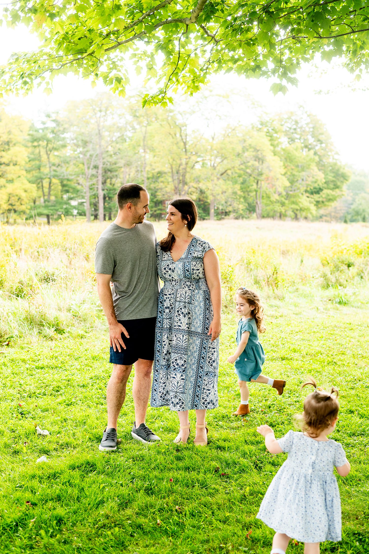 A happy family of four enjoys a sunny day outdoors, with parents smiling at each other while two young girls play nearby.