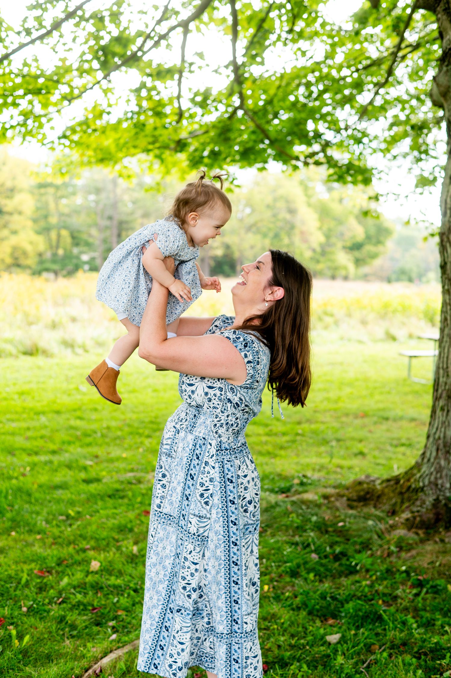 A mother in a blue floral dress lifts her smiling toddler daughter under a green tree in a sunny park.
