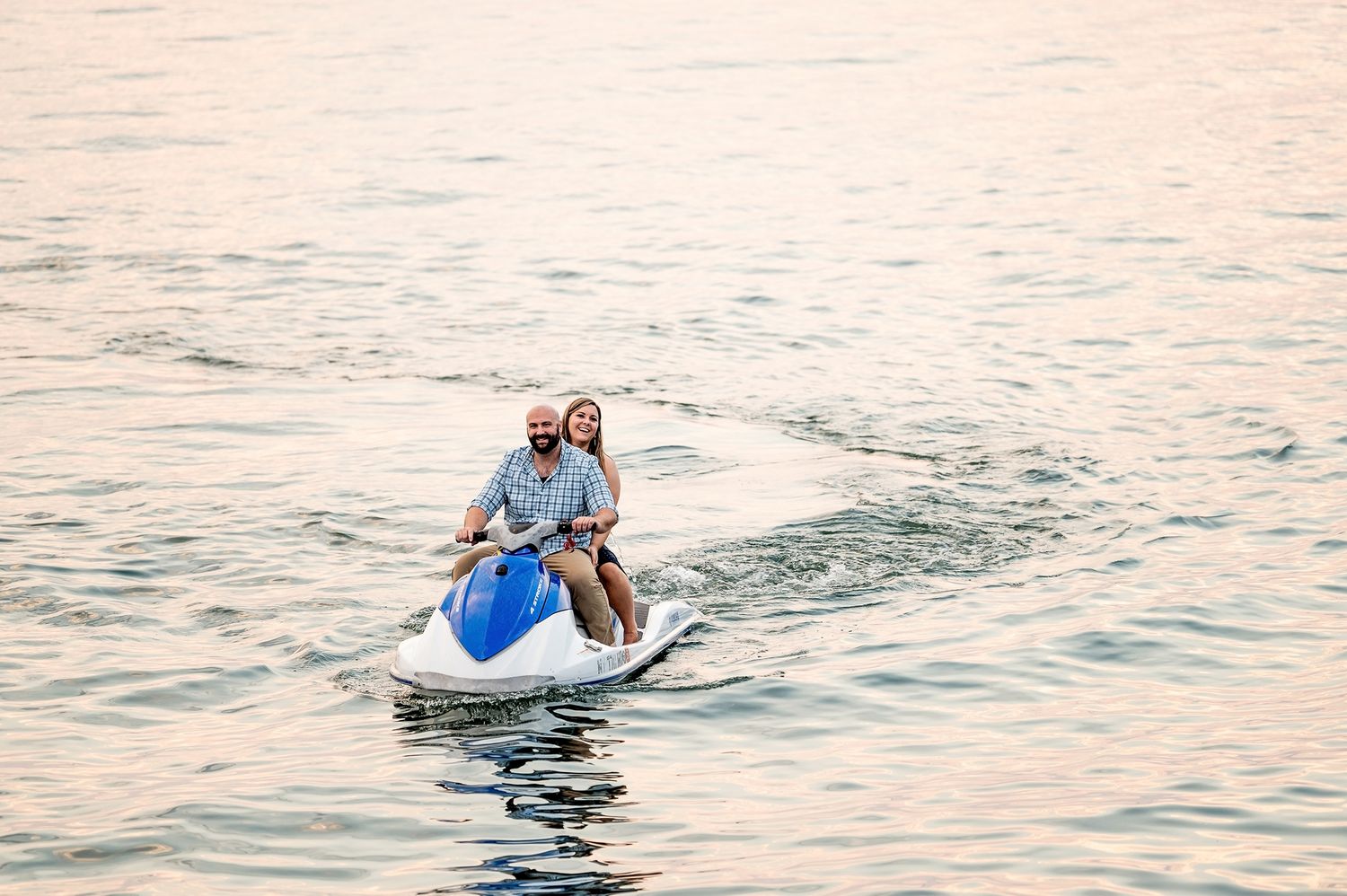 Couple smiling and riding a jet ski together on calm water at sunset.
