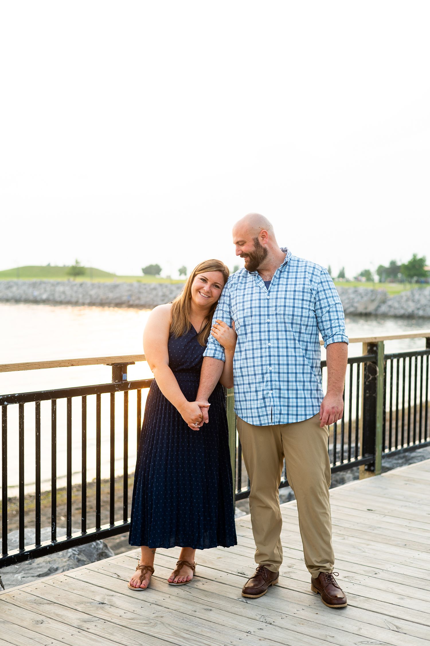 Couple posing on waterfront boardwalk, woman in navy dress and man in blue plaid shirt with khakis.