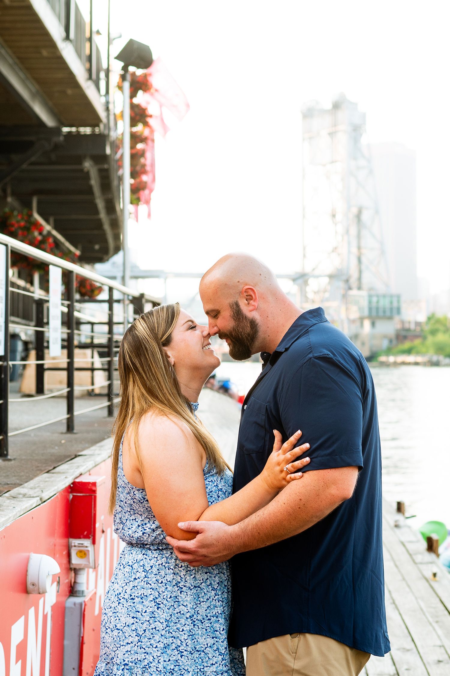 Couple sharing a romantic moment on a waterfront dock, woman in blue floral dress embracing bearded man in navy shirt.