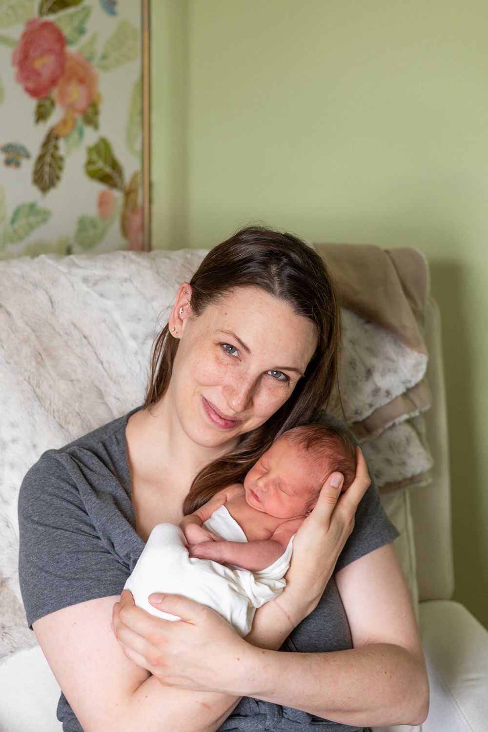 A tender moment between parent and newborn baby as they cuddle together on a cozy couch with floral wallpaper in background.