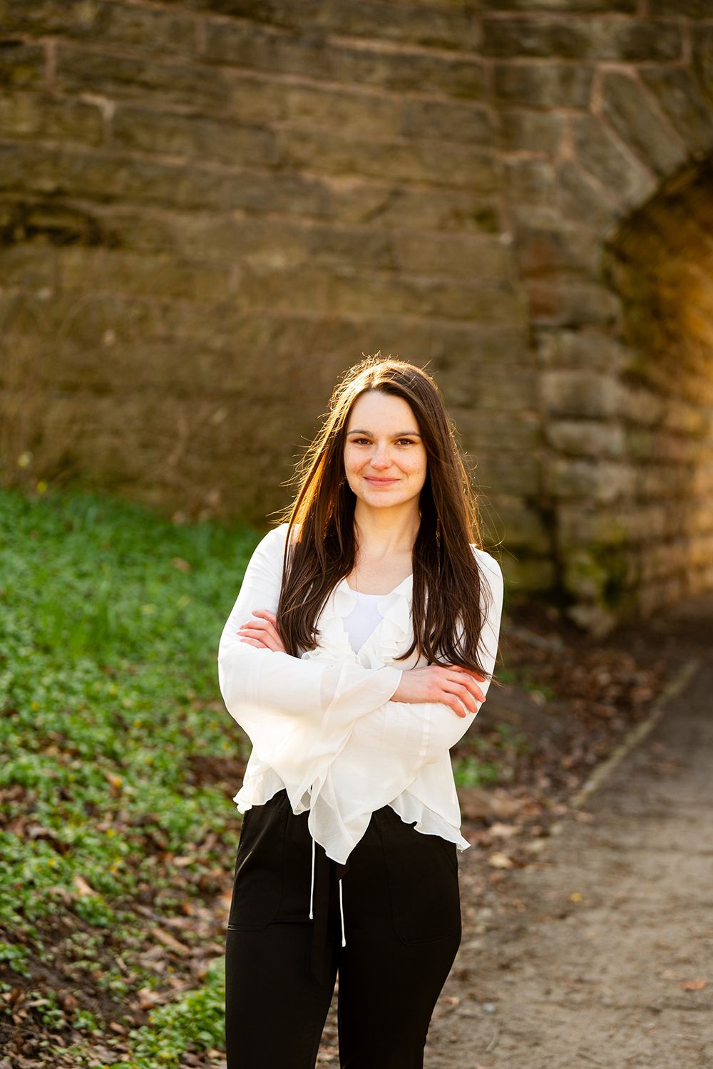 A person stands on a pathway wearing a white flowy top and black pants against a stone wall backdrop.