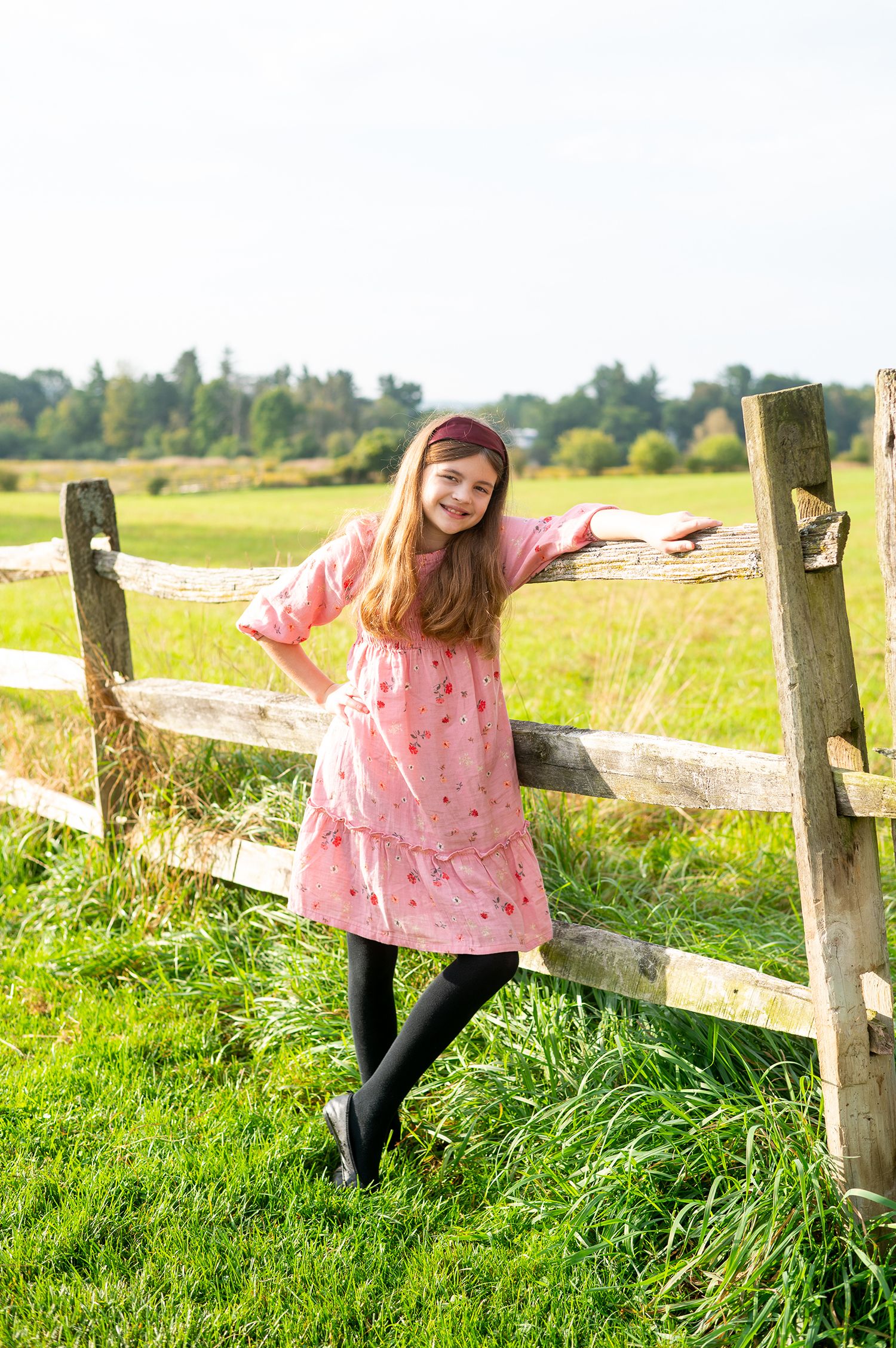A cheerful girl in a pink dress leans against a rustic wooden fence in a lush green countryside setting.